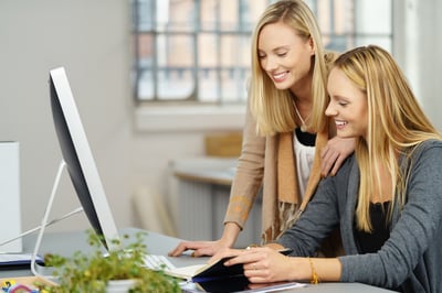 Two Young Office Women Reading Some Notes Together at the Table with Happy Facial Expressions.